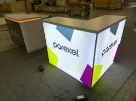 RENTAL: RE-1572 L-Shape Backlit Counter, RE-1576 White Laminated Counter with Recessed LED/RGB Lighting at Top Perimeter, SEG Backlit Fabric Graphics, and Vinyl Applied Graphic